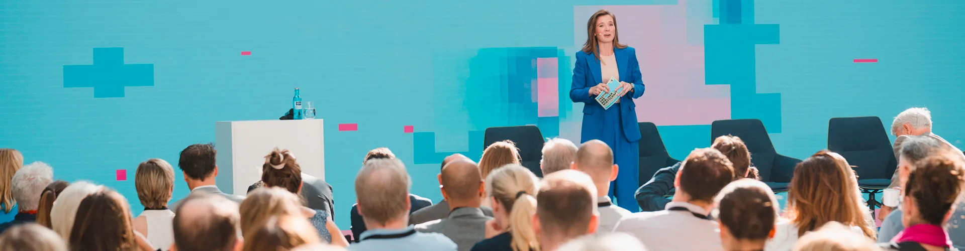 "Eine Frau in einem blauen Anzug hält vor einem Publikum eine Präsentation auf einer digitalen Bühne."