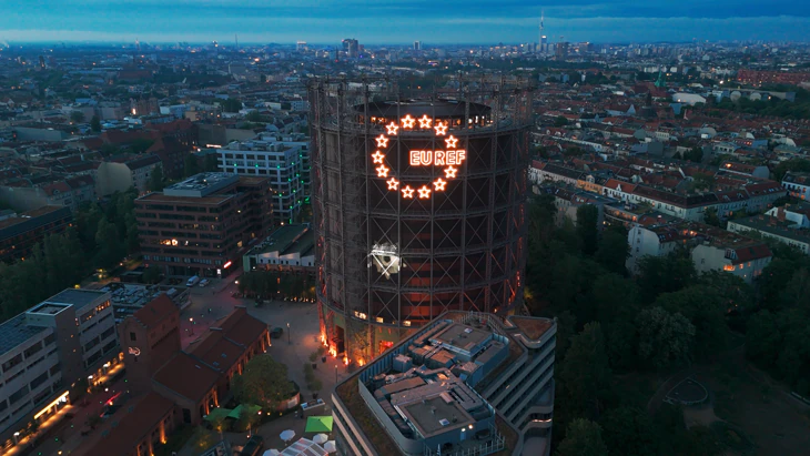 EUREF-Gasometer in Berlin bei Dämmerung mit leuchtendem Sternenkreis-Logo aus der Vogelperspektive.