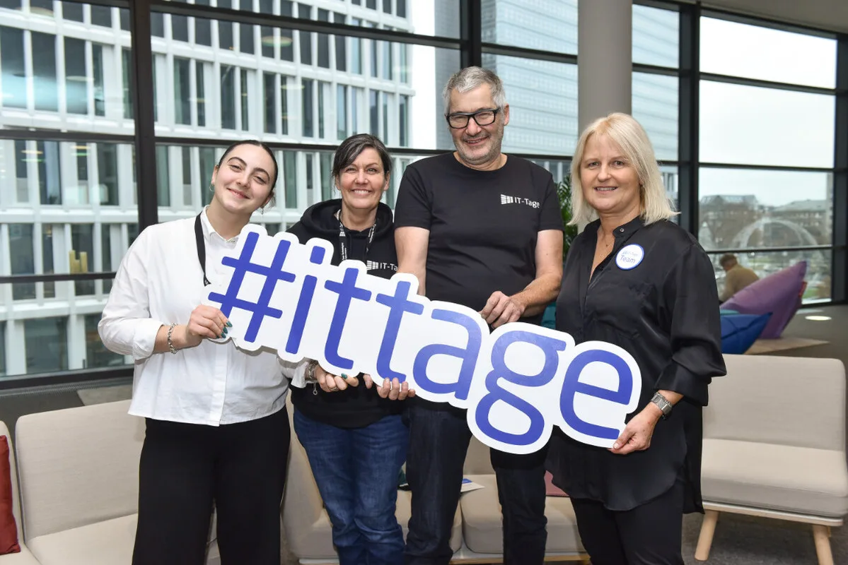 "Vier Personen halten gemeinsam ein großes „#ittage“-Schild in einem hellen, modernen Innenraum."
